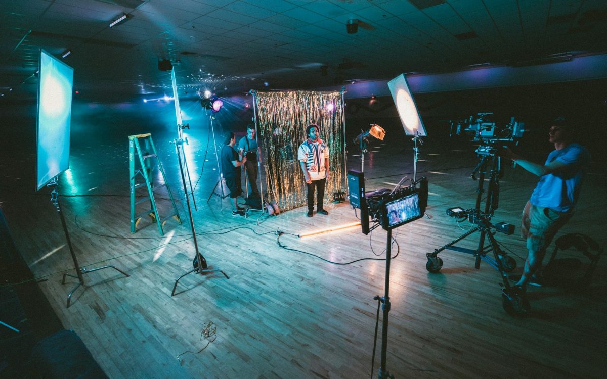 entry-level arts jobs. Photo: Jakob Owens, Unsplash. A figure stands in the centre of a dark indoor space with a photography set-up. Lets are shining on him and people are sorting out technical things with a camera.