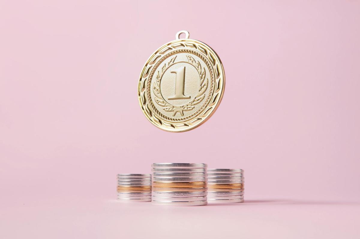 A pale pink background. There is a large gold coin with the number 1 on it hanging above three piles of coins.