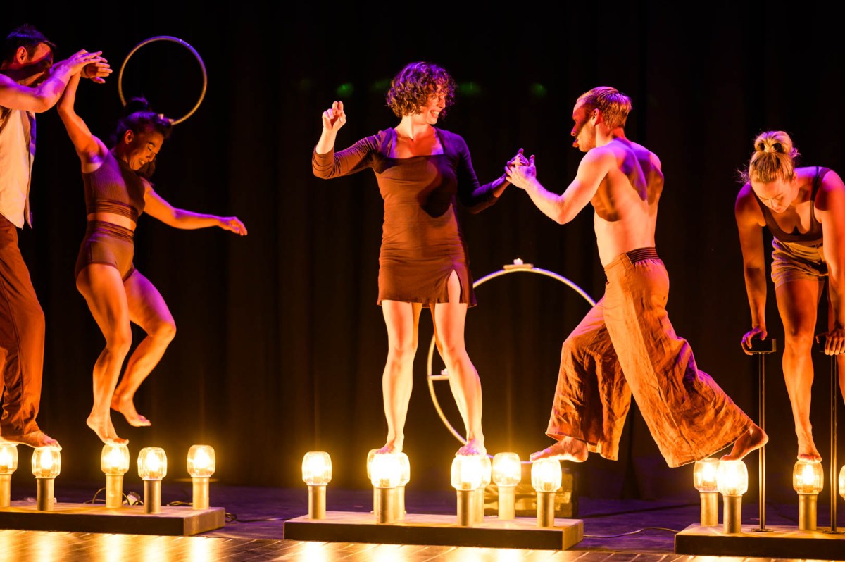 Five performers in shades of brown are walking on top of candle jars.