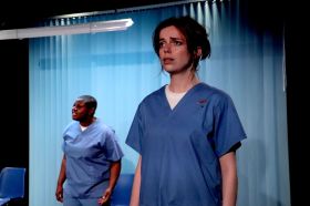 Two stressed-looking women wearing blue nurses uniforms stand on stage, with a light blue curtain in the background. The woman in the foreground is pale-skinned with short dark hair; the woman in the background is dark-skinned with very short black hair.