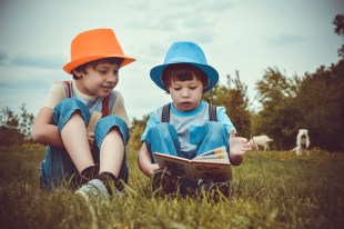 Two young boys are looking through a picture book in a field of grass. They are seated. The left one has an orange hat, the right one a blue one.