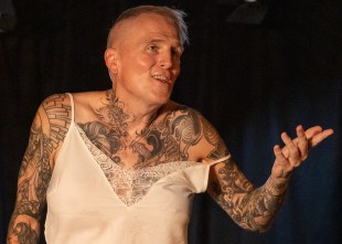 A trans woman in a white lace top. She is covered in tattoos and has short cropped fair hair.