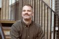 Sydney Festival's incoming Festival Director Kris Nelson. Kris sits on a stairwell smiling; he has short-cropped brown hair and a moustache, and wears an olive-coloured shirt.