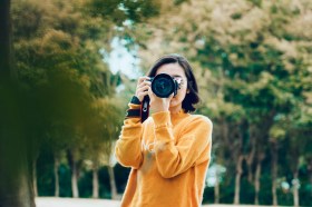 A woman with black hair and a yellow top has a camera covering her face. She is standing in front of a forest. Author photography headshot.