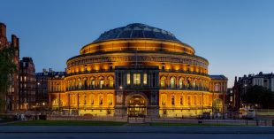 At night time a grand circular building with at least four storeys and a dome on top is lit up. The Royal Albert Hall.