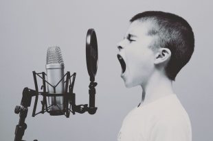 arts marketers. Image is a side on black and white shot of a young boy shouting with a very wide open mouth into a microphone that has a small screen in front of it. He is wearing a white T shirt and has close cropped hair.