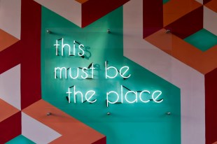 Neon sign saying "this must be the place" against geometric painted wall. Curator job