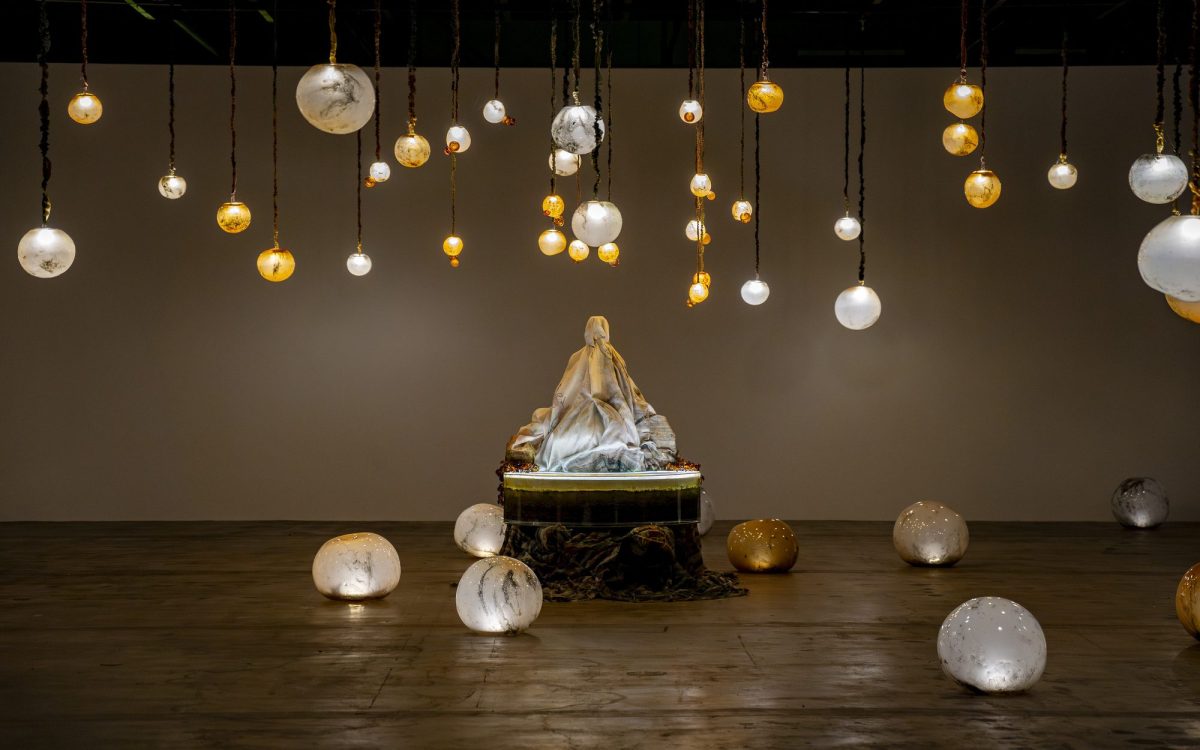 Marguerite Humeau, ‘*stir’, 2024, installation view at 15th Gwangju Biennale. Glowing round orbs are situated on the ground and hung from the ceiling around a statue-like figure covered by a white cloth.