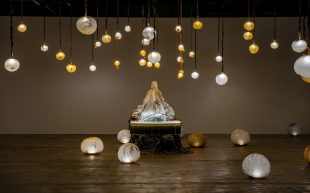 Marguerite Humeau, ‘*stir’, 2024, installation view at 15th Gwangju Biennale. Glowing round orbs are situated on the ground and hung from the ceiling around a statue-like figure covered by a white cloth.