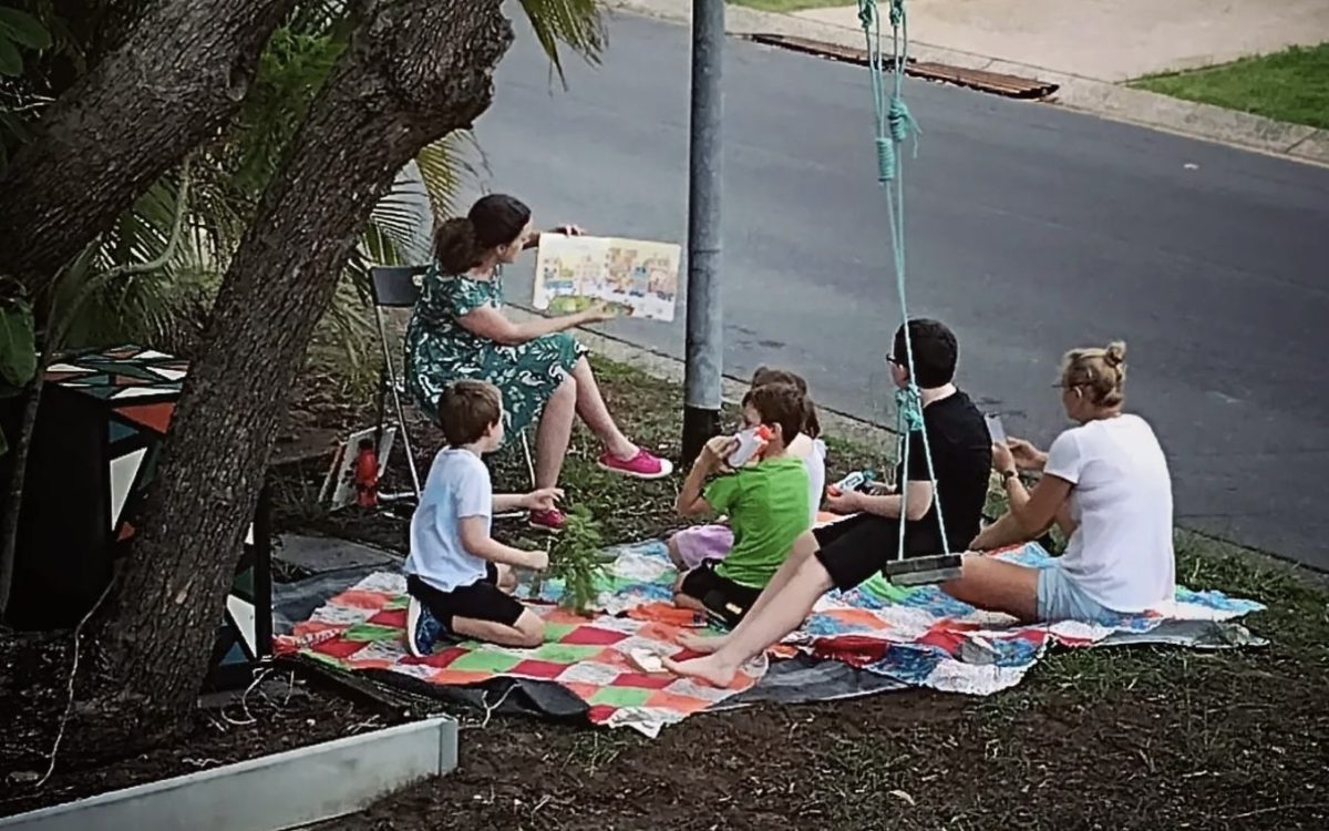 A woman is reading to children. She is sitting on a chair, they are on picnic mats. They are outside, under the shade of a tree.