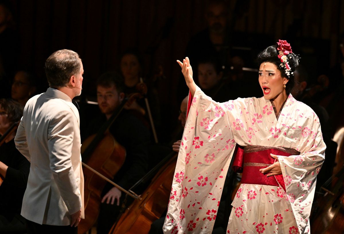 In the production of 'M. Butterfly', a singer, Kangmin Justin Kim, is dressed in a red and white kimono. She is facing Mark Stone, who is wearing a white jacket suit.