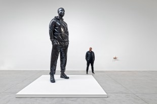 Over sized sculpture of an everyday man in white gallery, with artist dressed same as sculpture. Thomas J Price