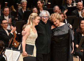 a grey haired conductor Michael Tilson Thomas hugs two female singers Siobhan Stagg (in a white dress), and Alice Coote (in a black dress), standing in front of an orchestra