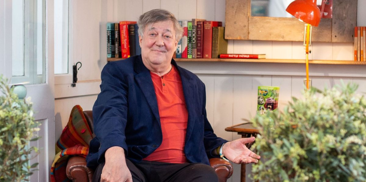 Stephen Fry at the Hay Festival of Literature and Arts, held annually in the booktown of Hay-on-Wye, Wales. A grey-haired, slightly plump Englishman smiles benignly at the camera. He wear a blue jacket over a red shirt and is sitting in a leather armchair. His right hand is on his knee; his left hand is turned upwards, as if encouraging comment or conversation.