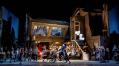 A street scene in the production of 'La bohème'. In the foreground is a man on a bicycle carrying some white balloons. There are crowds of people around him. English National Opera production.