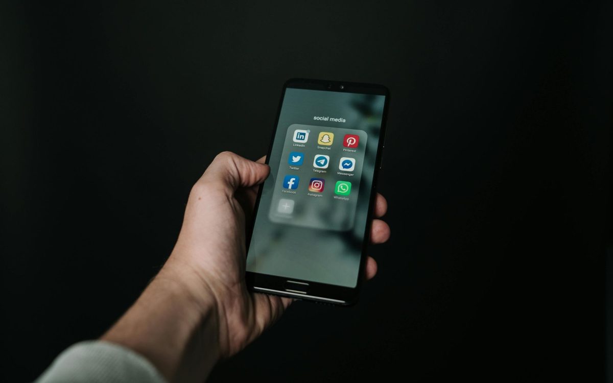 social media hacking: The hand of a white male person holding a smartphone displaying a range of social media icons on its screen against a black background.