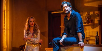 Lily Collins in a glittering outfit is standing and holding onto a glass of wine. She has blonde hair. Álvaro Morte, a dark haired, bearded man in dark clothing is sitting on a kitchen bench in a production of 'Barcelona.'