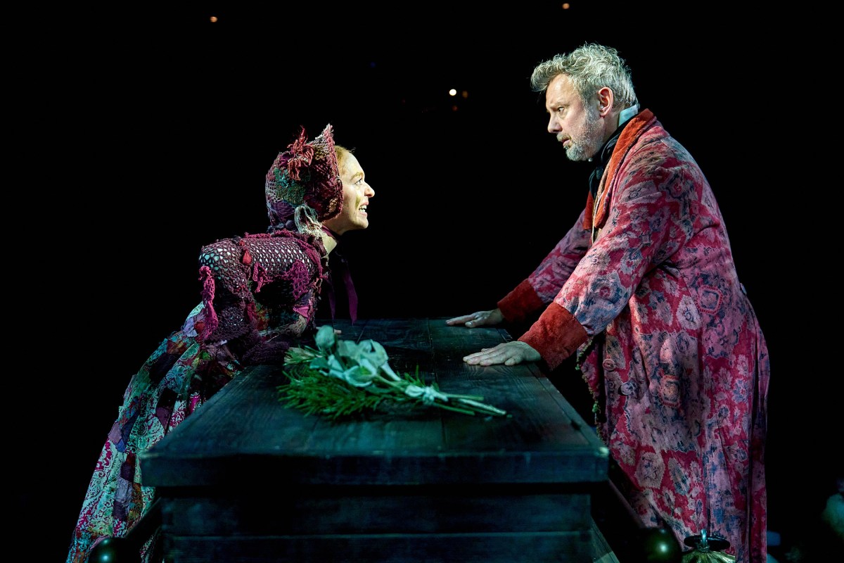 A woman in Victorian clothes, including a bonnet, leans over a table with a bunch of flowers on it towards a man in a Victorian dressing gown, who looks concerned. A Christmas Carol at the Old Vic