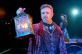 a man in Victorian clothes holds up a lantern with a grim look on his face. A woman in a bonnet and Victorian clothes stands behind him out of focus. A Christmas Carol at the Old Vic