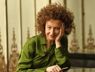 So you want my arts job? is ArtsHub's ongoing series exploring a wide range of arts careers. Pictured is Spanish conductor Conductor Lucía Marín, who has curly auburn hair, pale skin, and sits with her face resting in the palm of her left hand. She is wearing a green silk shirt; ornate wallpaper can be seen in the background.