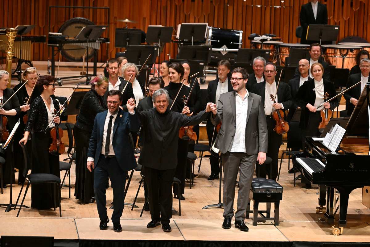 Three white men in suits raise their joined hands to take the applause in front of a standing orchestra. LSO