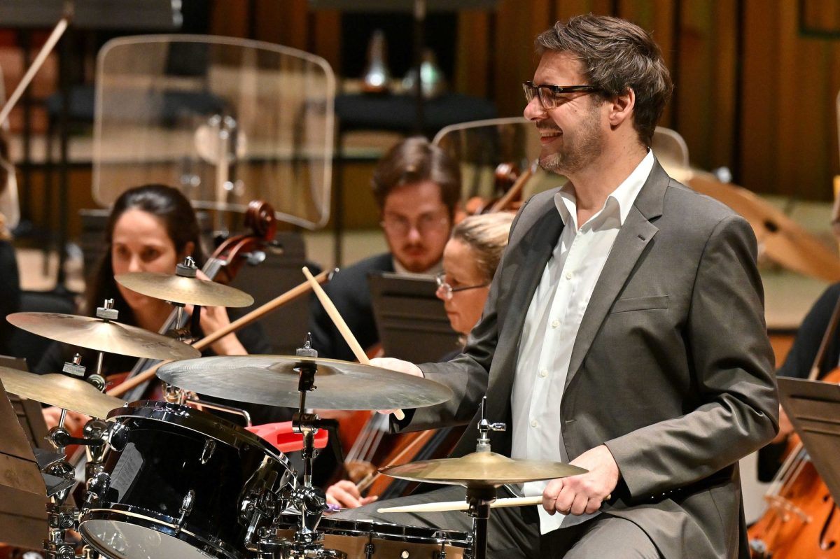 A smiling Caucasian man in a grey jacket, white shirt and glasses plays the drums in front of an orchestra and looks of to the left of the frame. LSO