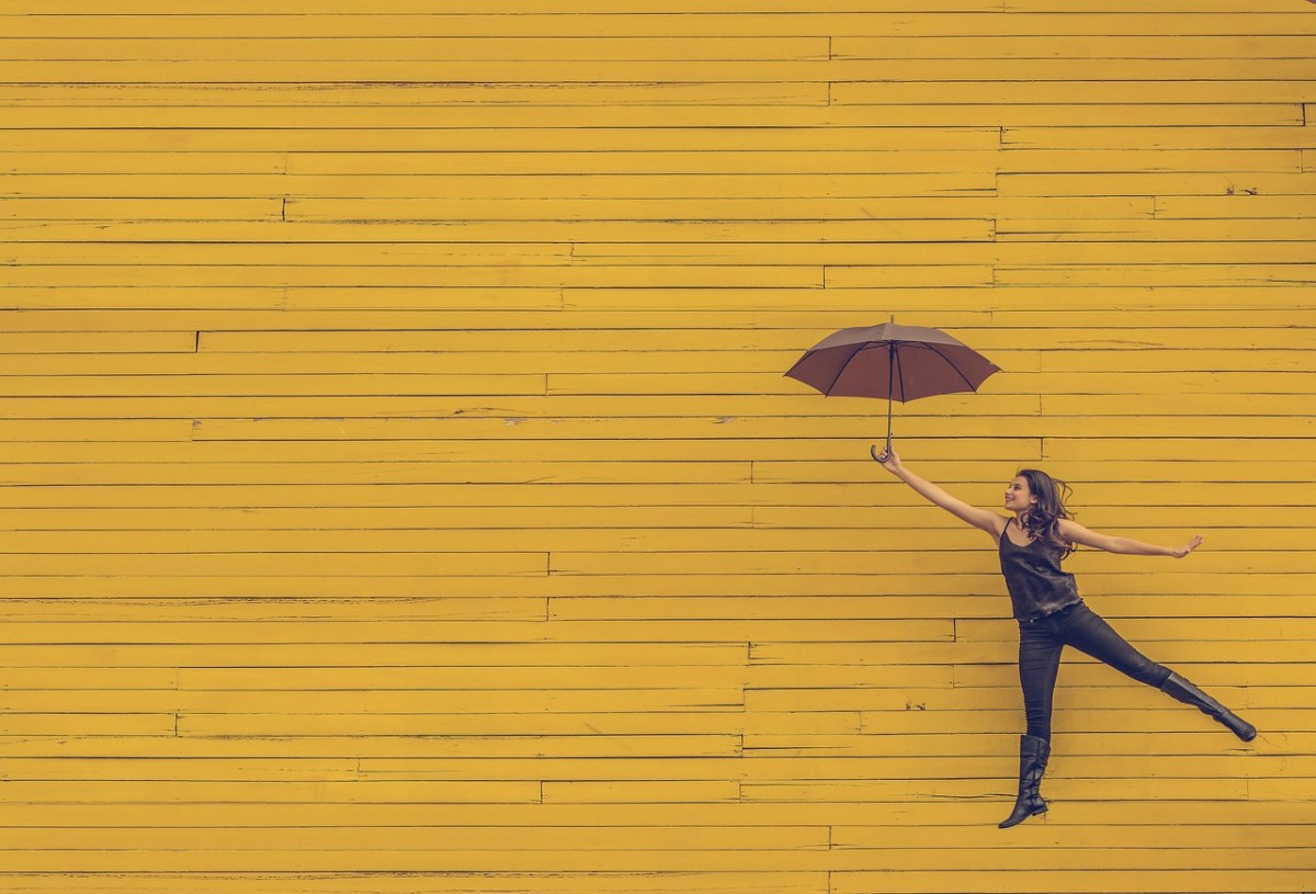 Against a yellow background, a woman is floating and carrying an umbrella.