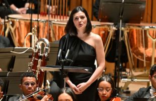 French mezzo-soprano Marianne Crebassa sung with beautiful pitch and gentle emotionality in her appearance as soloist with the Simón Bolívar Symphony Orchestra of Venezuela. The photograph shows a slender, pale-skinned woman with shoulder length black hair and wearing a black dress that reveals her left shoulder. She stands to sing, surrounded by seated members of the orchestra.